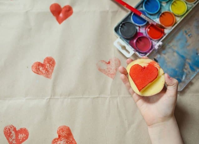 A child's hand stamps a heart onto a piece of fabric. The stamp is made of potatoes. There's a palette of water colors next to the hand as well.