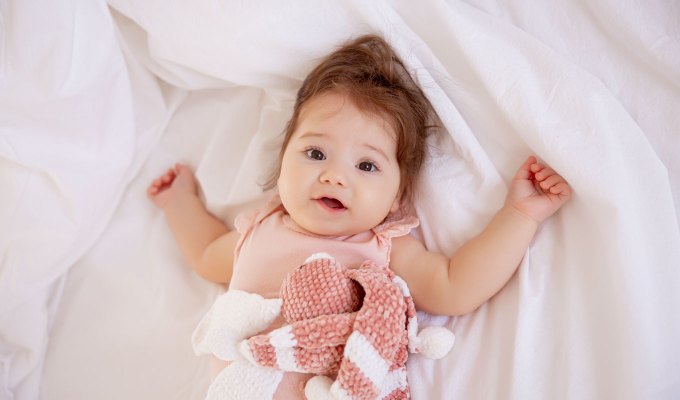 baby-names-that-mean-love: Baby girl in pink bodysuit lying on the bed at home looking at the camera, top view.