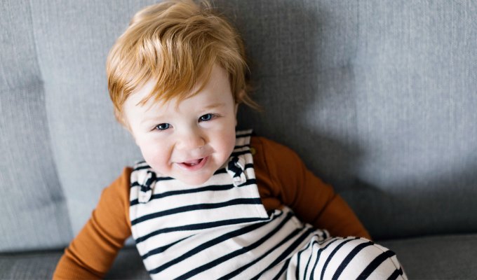 baby-names-that-mean-love: A young white boy with ginger hair sits on a couch and smiles at the camera. He wears an orange long sleeved top and black and white striped overalls. The couch is gray.