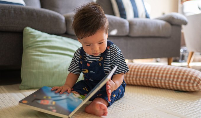 baby-names-that-mean-love: Baby boy looking at a book in the living room sitting on a baby safe soft play mat on the floor at home. He seems to be mixed race with brown hair and brown eyes. He wears a striped shirt and overalls with little designs.