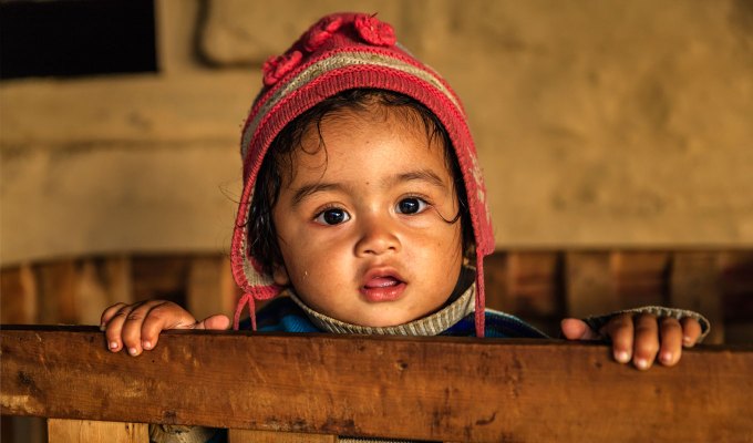 baby-names-that-mean-love: A young Nepali boy holds onto the railing of a play pen. He looks at the camera. He is wearing a red winter hat with flaps.