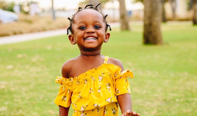 baby-names-that-mean-love: A young Black girl with hair twists wears a yellow romper while running around outside in a lush green park. She is smiling.