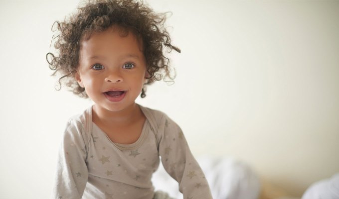 baby-names-that-mean-love: A photo of a young girl in her pajamas sitting on a bed. She seems to be mixed race and has brown curly hair and light eyes. Her pajamas are a light gray with stars.