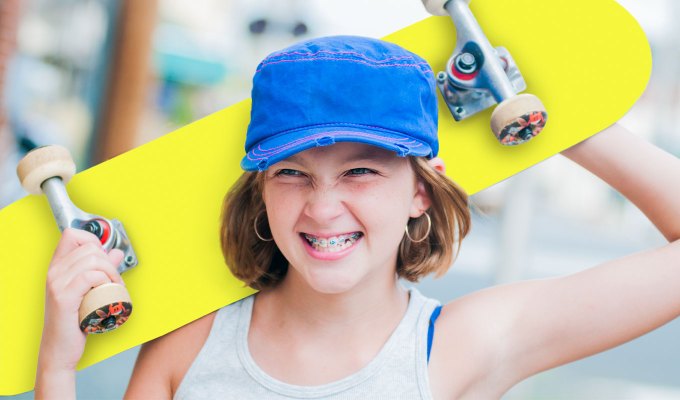 baby-names-that-mean-love: A young girl with tomboyish style raises a skateboard above her head. She is white and has brown hair and braces. She wears a white tank top and a blue hat.