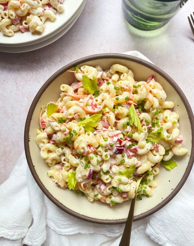 big batch recipes: bowl of classic macaroni salad with a spoon it it, on a table next to a glass and stack of plates