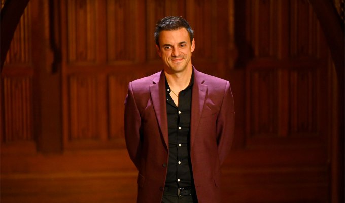 the-traitors-season-2-cast-ranked: A photo of Dan Gheesling standing in front of the castle in a maroon suit and black top.