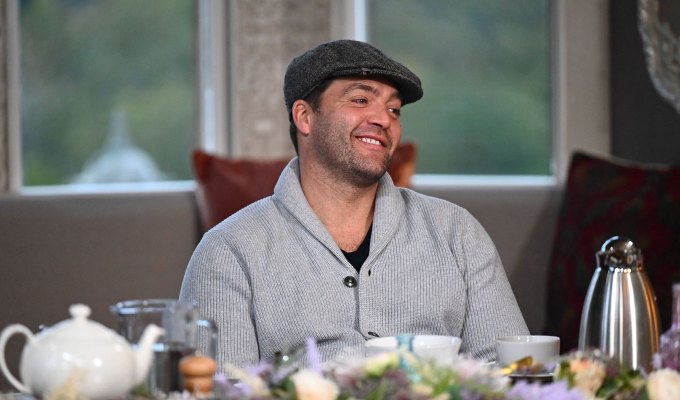 the-traitors-season-2-cast-ranked: A photo of Chris C.T. Tamburello sitting at the breakfast table wearing a gray sweater and a golfer hat.